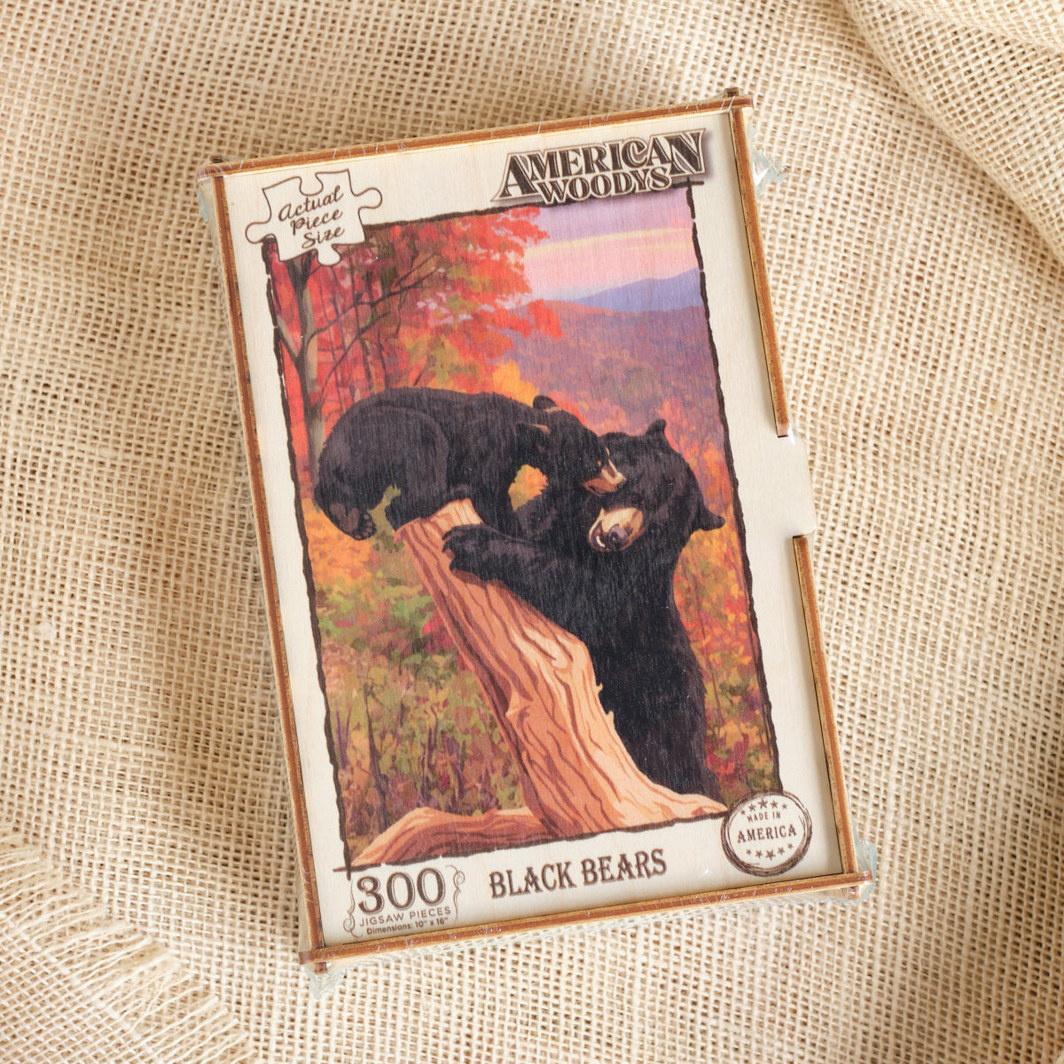 A boxed jigsaw puzzle titled "American Woods" featuring an illustration of two black bears on a tree trunk in an autumn landscape. The box is placed on a textured beige fabric background.