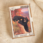 A boxed jigsaw puzzle titled "American Woods" featuring an illustration of two black bears on a tree trunk in an autumn landscape. The box is placed on a textured beige fabric background.