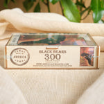 A boxed jigsaw puzzle labeled 'BLACK BEARS 300 JIGSAW PIECES' sits on a beige fabric surface with blurred green leaves in the background. The box shows a rustic wood-toned design with a small bear illustration and 'Made in America' badge.